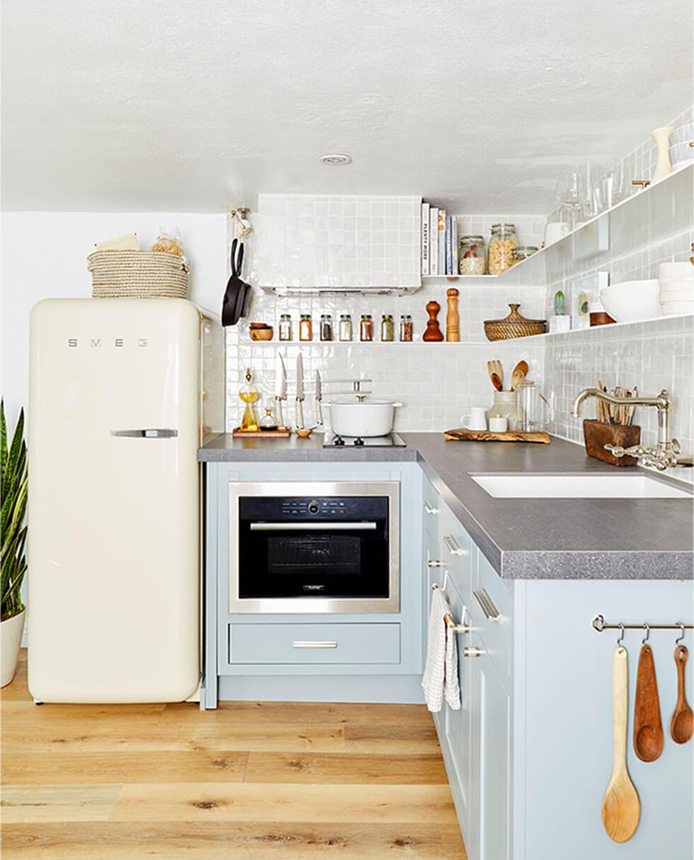 Small kitchen with wooden floors, white Smeg fridge, light blue cabinets, and lots of racks, shelves, and hanging spoons and tools.