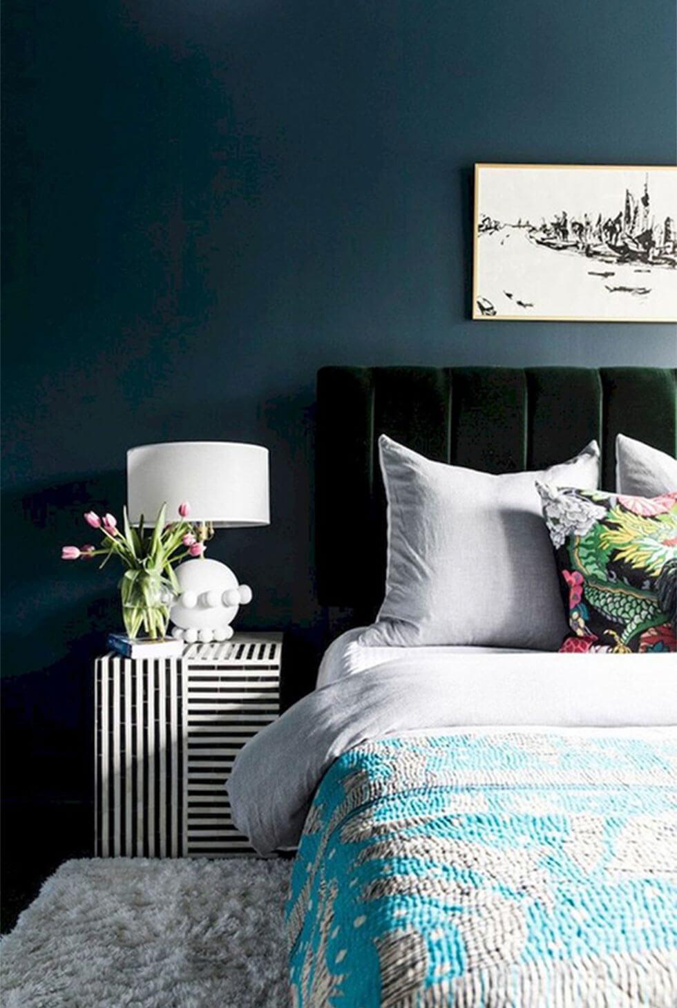 A bedroom wall painted in deep navy blue with an emerald velvet headboard and light coloured bedding.