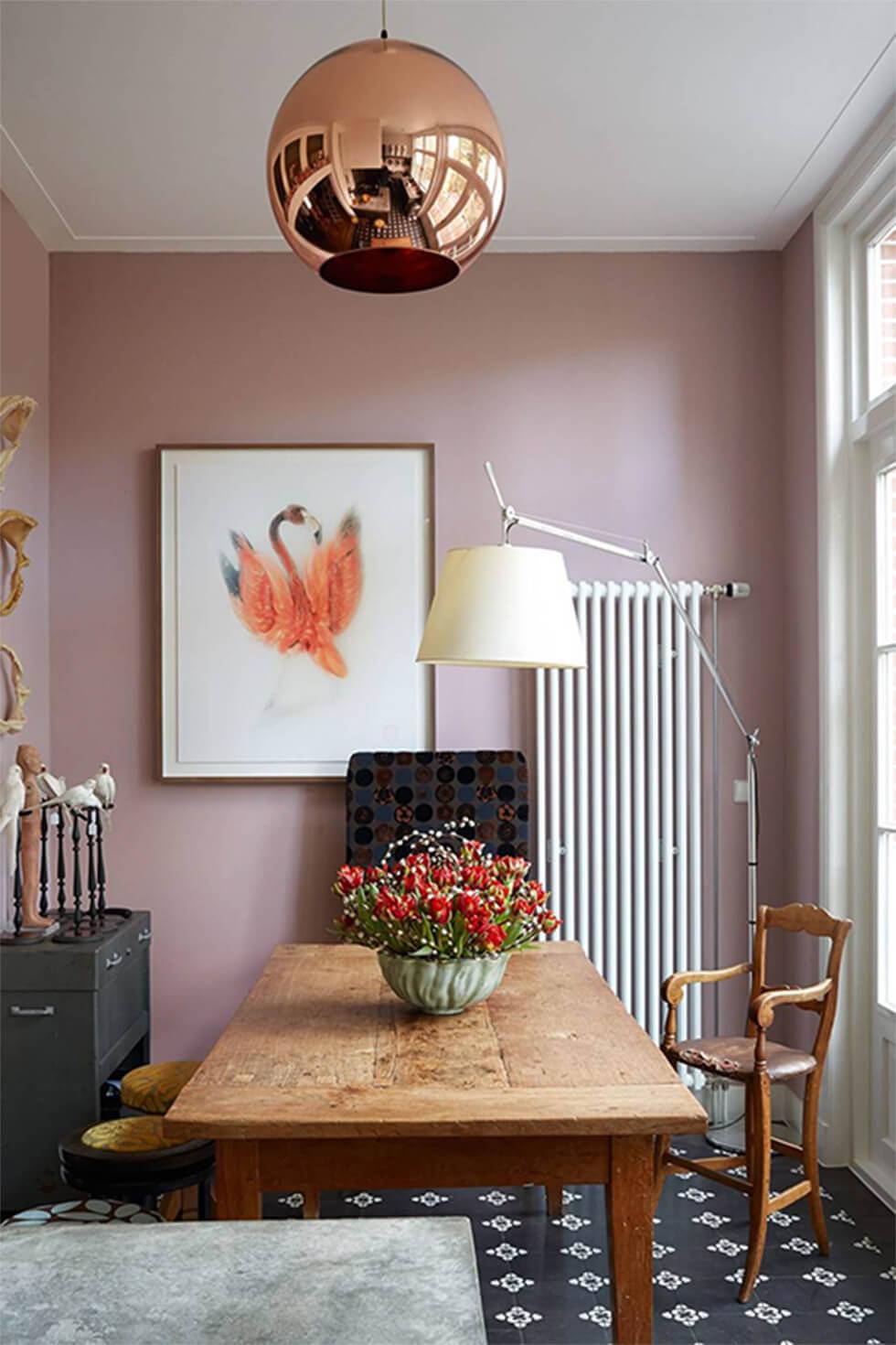 Eclectic small dining room with narrow rectangular wooden table, rose gold lamp, soft mauve walls and framed artwork