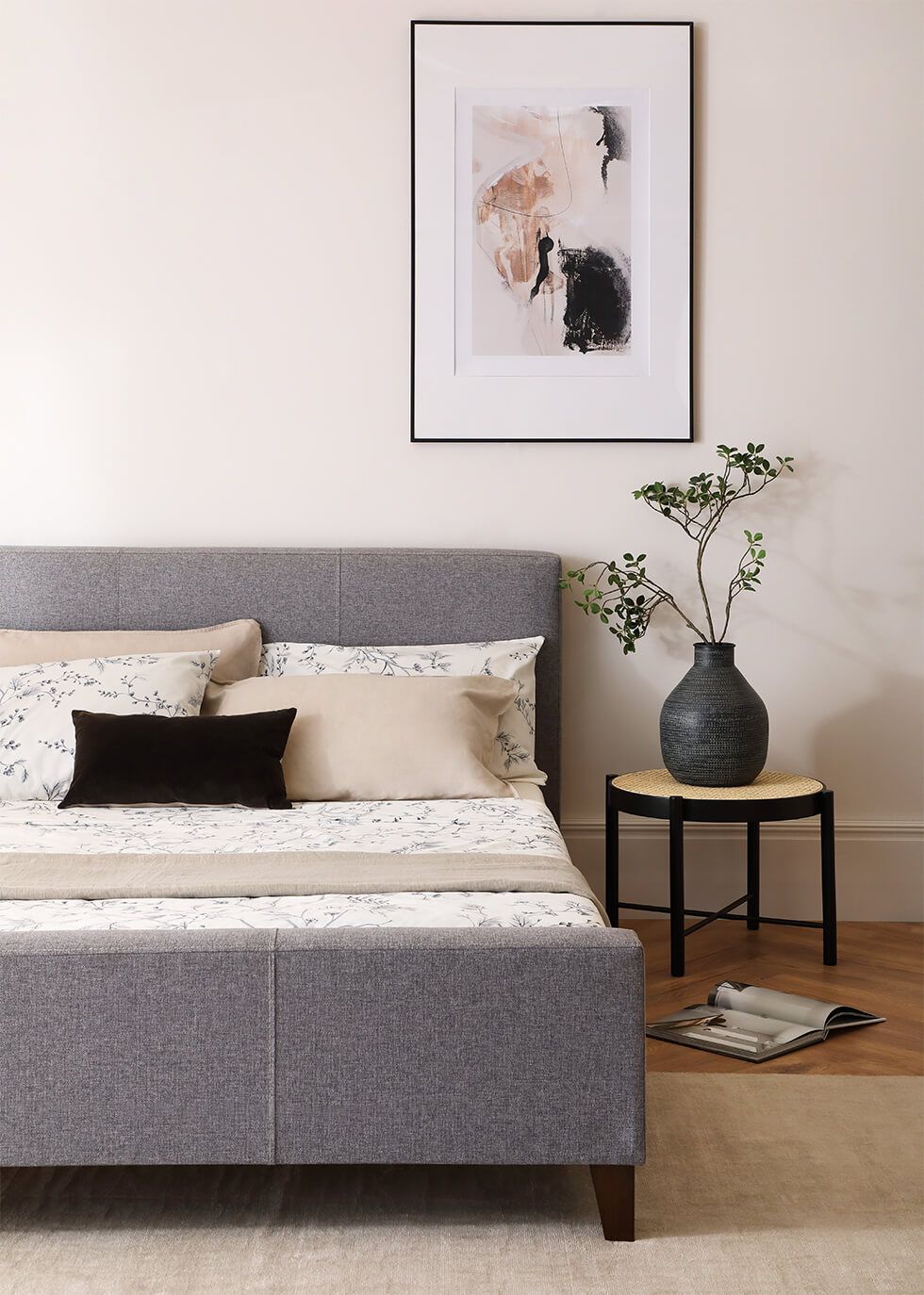A minimalist bedroom with a fabric grey bed, black side table and abstract wall art