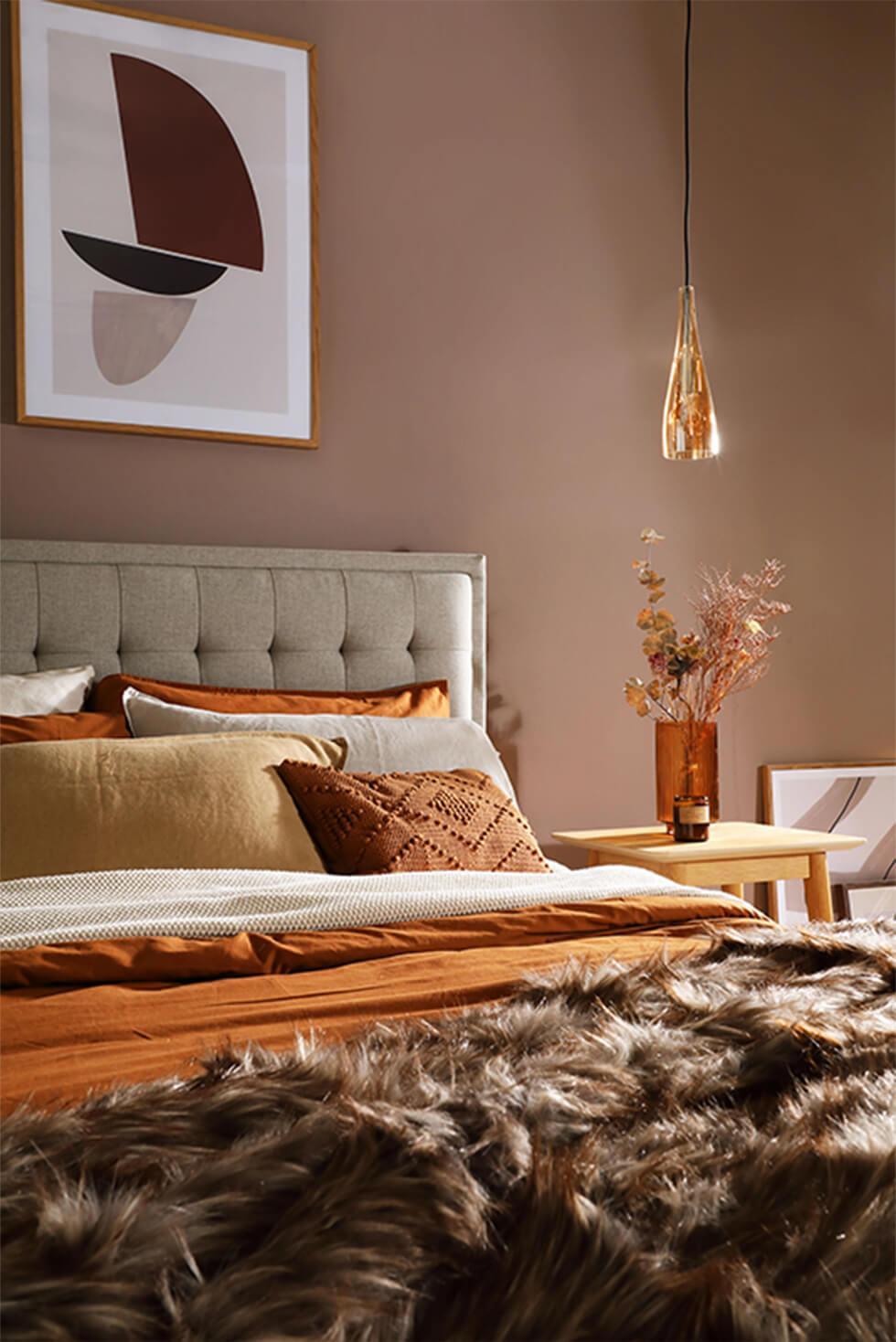 Warm glamorous bedroom with beige fabric bed, orange and brown bedding, fur throw and brass lighting
