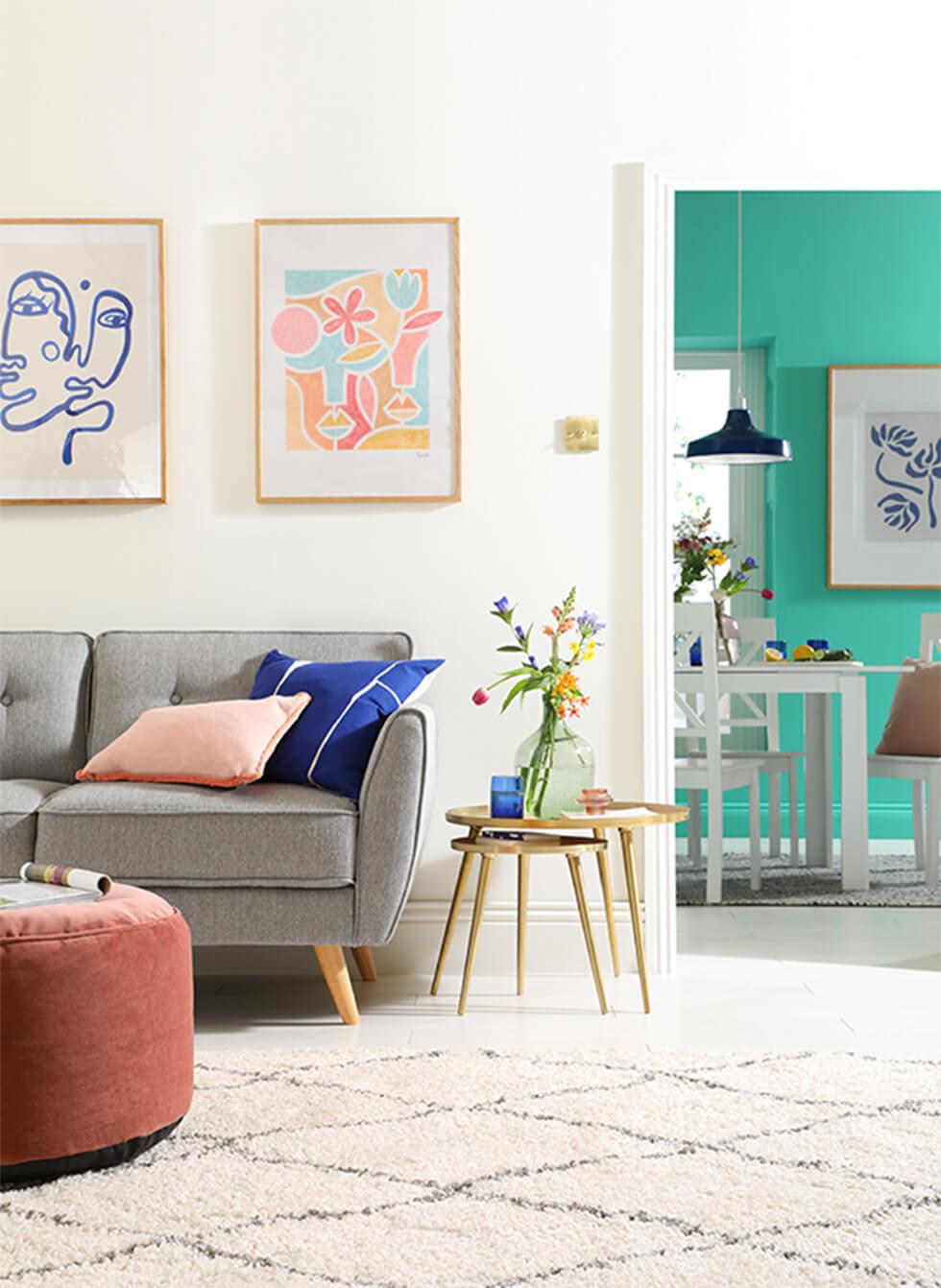 Neutral living room with grey sofa, velvet pouffe, pink and blue cushions and flowers, with a view to a turquoise dining room