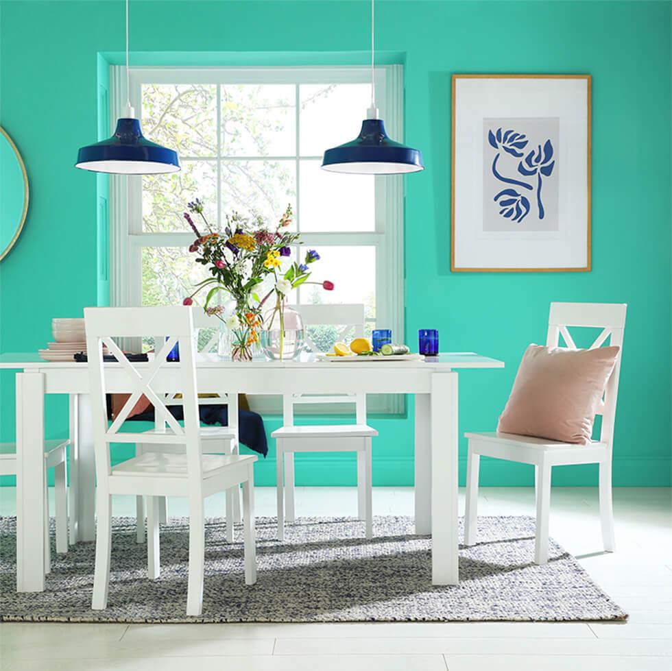 Pastel turquoise dining room with white wooden dining set, blue lamps and glassware, pink cushion and a grey rug