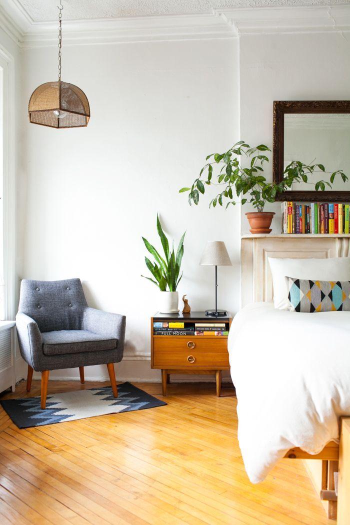 A light and airy bedroom with wooden floors, white bedding, a grey armchair and plants.