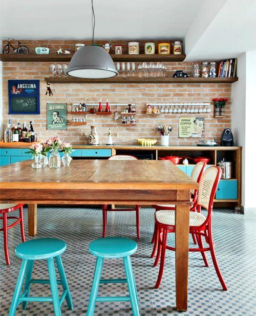 A dining room and kitchen with a large wooden table, tiled floors, brick walls and colourful chairs.