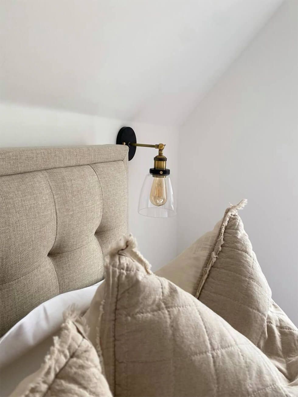 A beige tufted bed with comfy pillows next to a vintage wall sconce