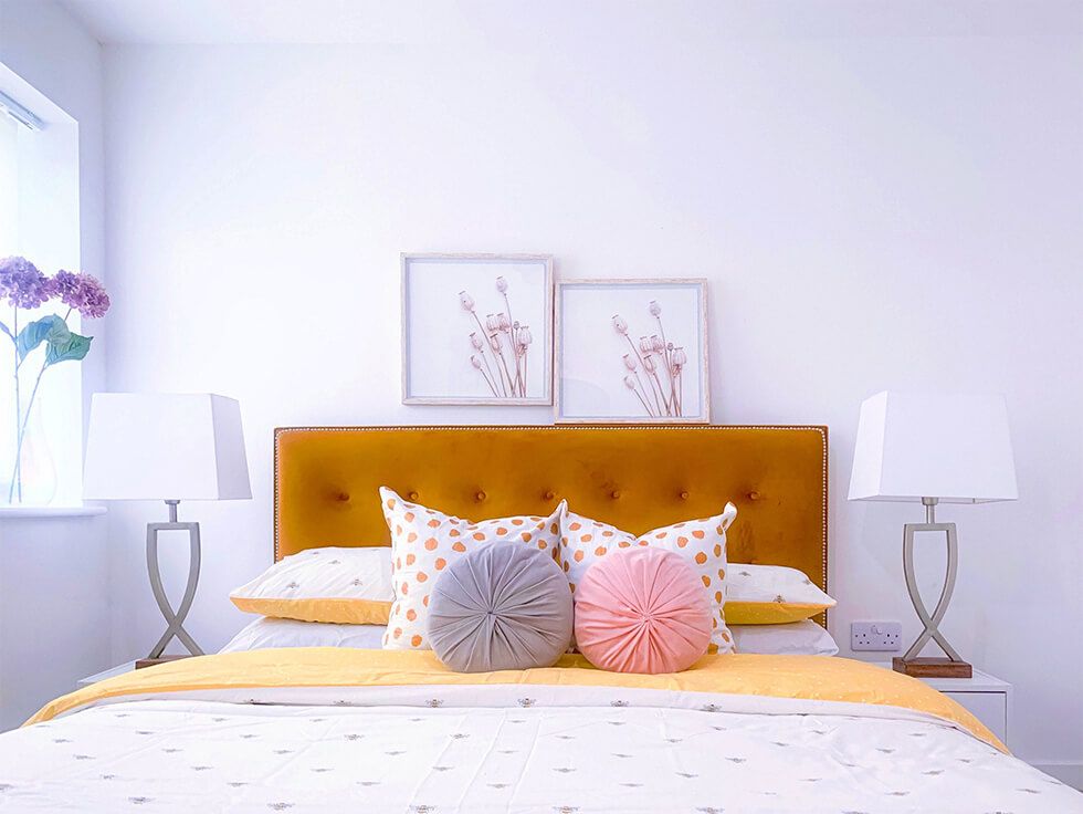 All white bedroom centred with a mustard bed and decorated with colourful pillows