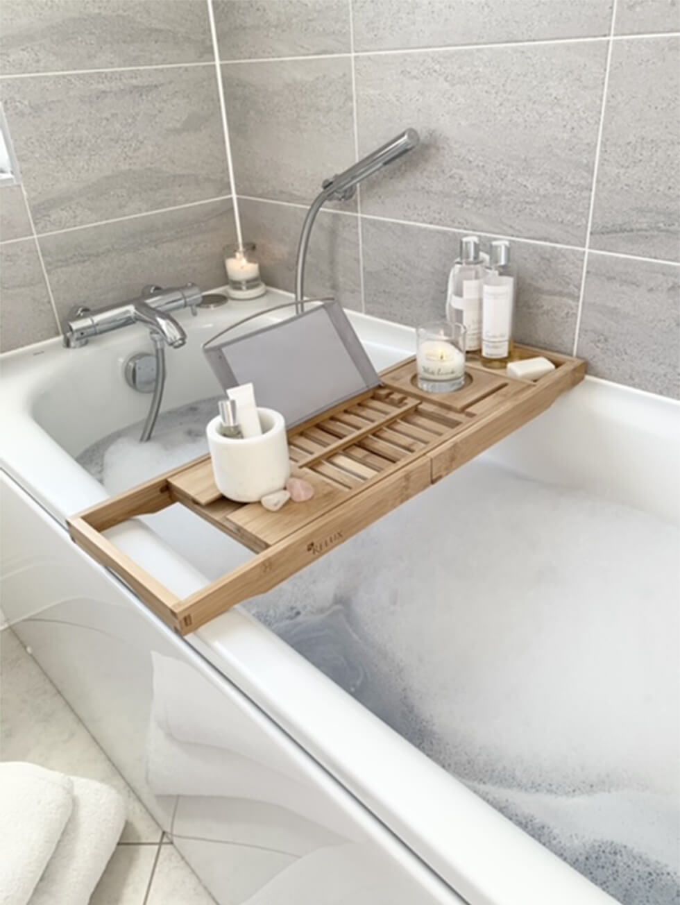 A bubble bath with a wooden bath tray decked with toiletries and a scented candle