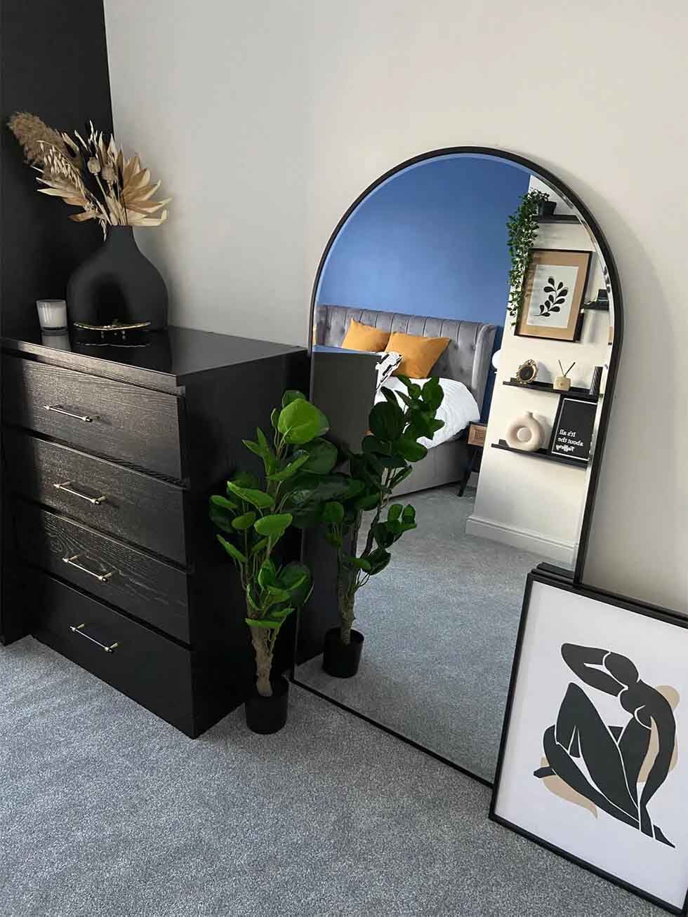 A black chest of drawers next to an arched mirror and a framed abstract art piece