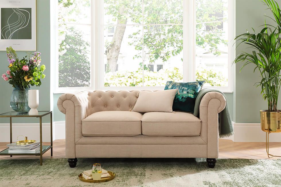 A sunny and relaxing living room with an oatmeal fabric Chesterfield sofa, indoor plants and fresh flowers