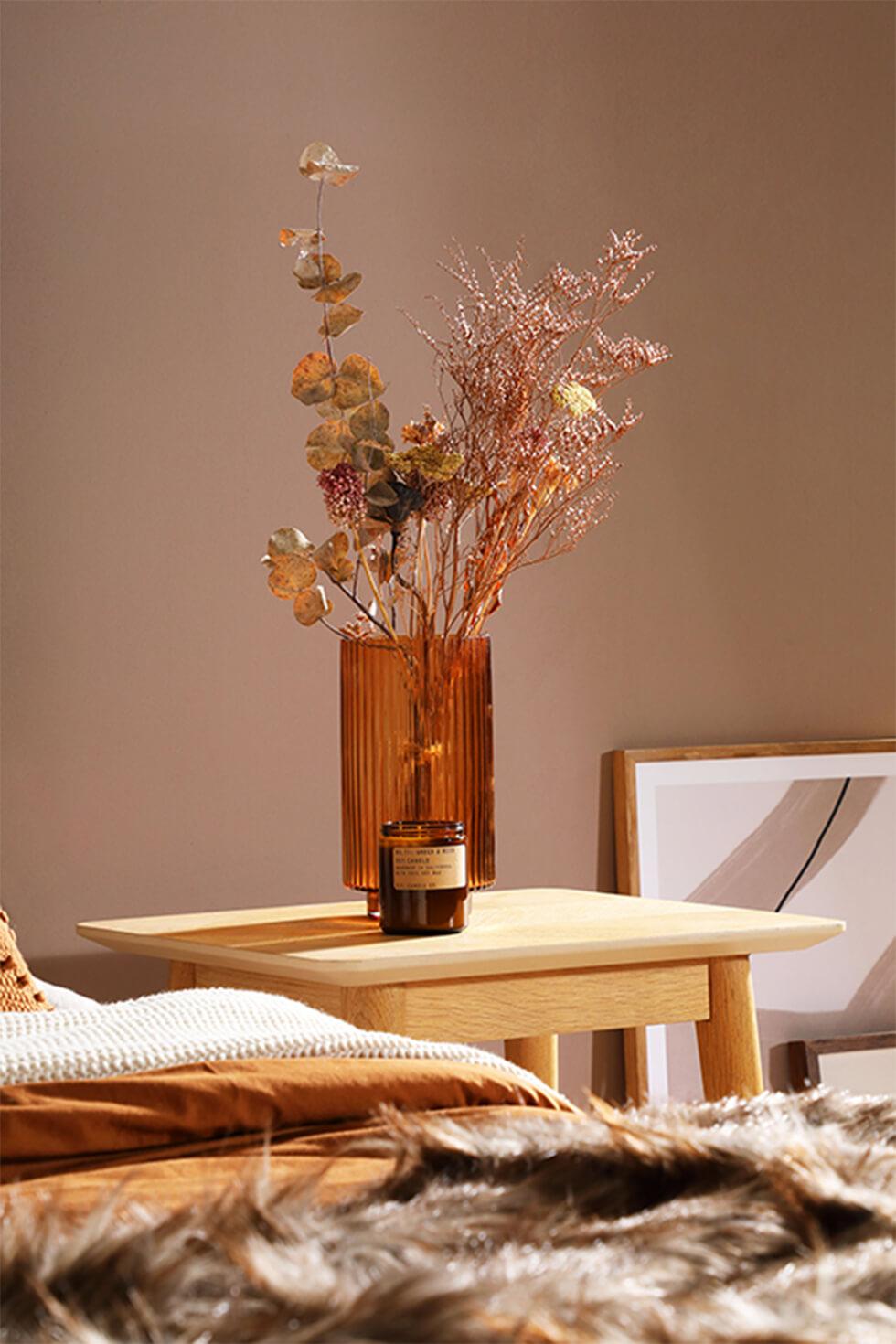 Warm toned room with wooden bedside table, brown glass jar with dried flowers and fur throw